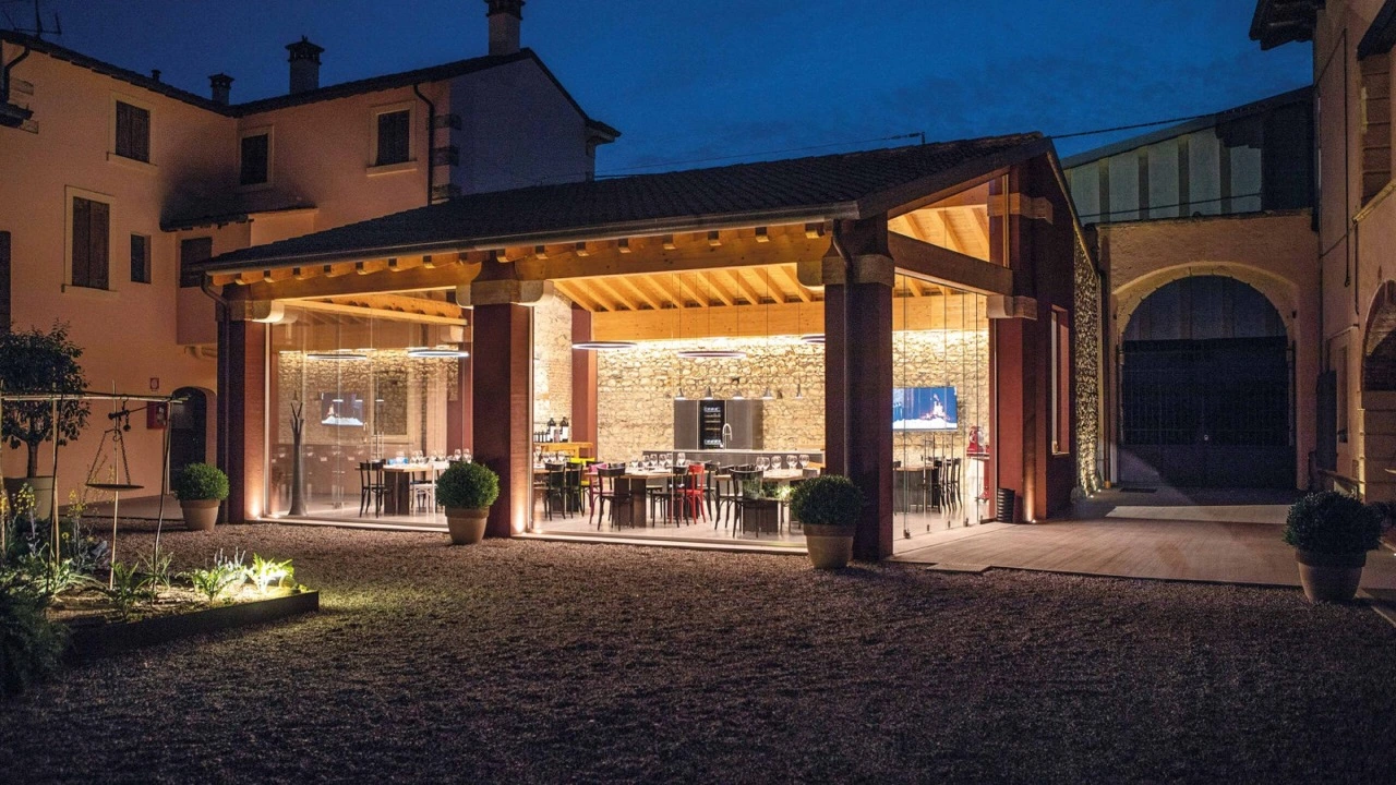 Vista notturna della Wine Boutique di Cantina Farina a Pedemonte di San Pietro in Cariano, con struttura in legno e vetro illuminata, tavoli apparecchiati per la degustazione e corte storica sullo sfondo, nel cuore della Valpolicella Classica